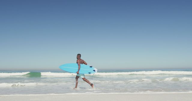 Happy african american man carrying surfboard running on sunny beach in the sea. Summer, hobbies, surfing and vacations.