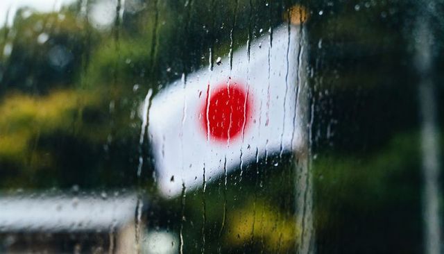Rain-soaked japanese flag fluttering beyond window with rain streaks and blurred red disc