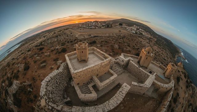Sunset-lit coastal fortress overlooking rugged cliffs and sea with watchtowers, courtyards