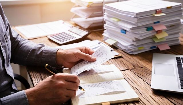 Hands comparing receipts while writing ledger on cluttered desk with laptop and invoices