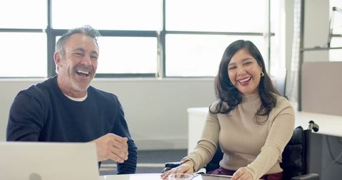 Diverse coworkers laughing and collaborating around desk with laptop and wheelchair in sunlit office