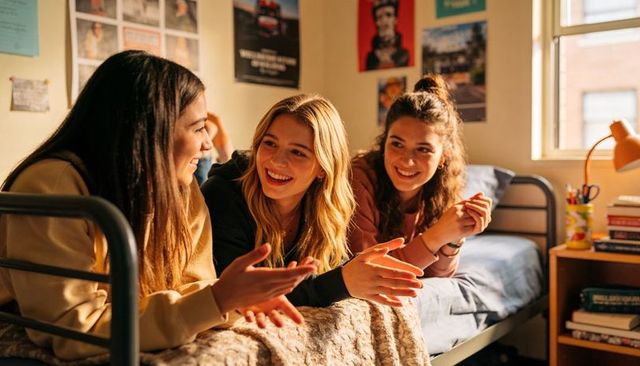 College roommates chatting on dorm bed in warm sunlight, studying and laughing together