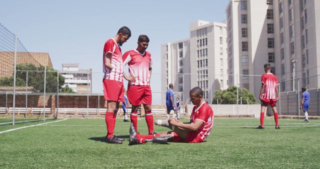 Three soccer players in red jerseys are on an urban soccer field. Two are standing while another is sitting on the ground, appearing to provide assistance to an injured teammate. Residential buildings are visible in the background. Useful for themes of sportsmanship, teamwork, urban sports, and injury recovery.