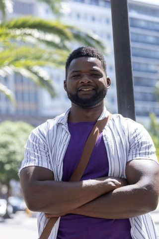 Confident african man standing with arms crossed in sunny urban plaza wearing purple shirt