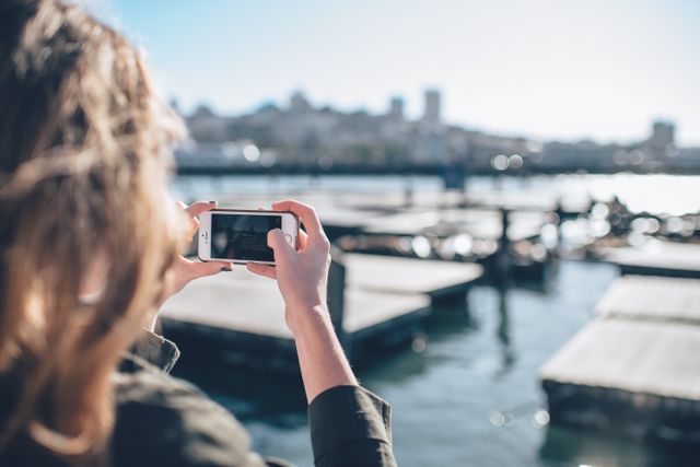 Person taking photo of cityscape with smartphone near waterfront. Might be used for content on tourism, urban exploration, outdoor activities, or promoting city attractions.