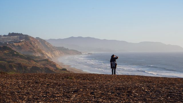 Person standing on cliff edge overlooking vast ocean and sandy beach with mountains in distance. Ideal for travel blogs, outdoor adventure themes, peaceful and relaxing getaway promotions, or nature photography collections.