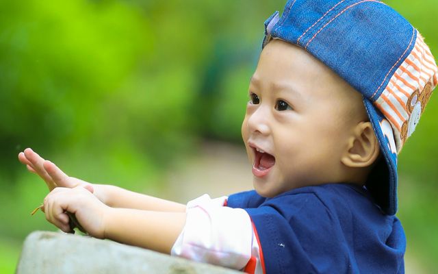A toddler joyfully plays outdoors while wearing a denim cap backward. The background features lush green foliage, suggesting a park setting. The child appears delighted, enhancing the image's joyful and happy mood. This image is ideal for use in advertisements, family-oriented blogs, children's product promotions, and articles focused on early childhood development and parenting.