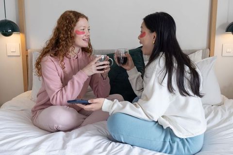 Two friends relaxing on bed in loungewear wearing under-eye gel patches sharing wine