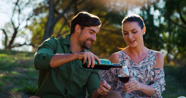A young Caucasian man pours wine into a glass held by a young Caucasian woman, both enjoying a moment outdoors, with copy space. Their relaxed and happy expressions suggest a romantic or celebratory occasion in a natural setting.