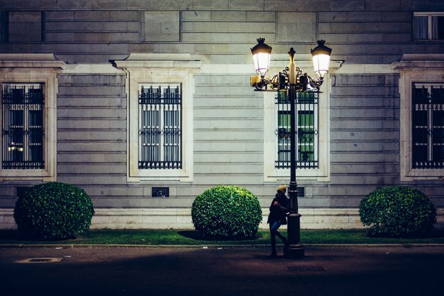 A woman stands alone by a lamp post in front of a historic building at night. The building features large windows with metal grills and neatly trimmed spherical bushes. This image can be used in projects related to urban living, solitude, historic architecture, and night photography.