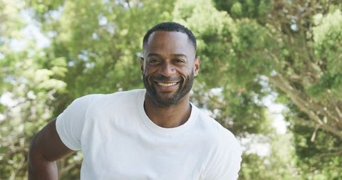 Smiling man leaning forward in white tee enjoying sunlit park with lush green trees