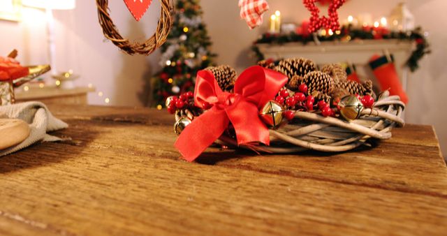 Wooden table decorated with a rustic Christmas wreath featuring pinecones, red ribbon, and bells. Background includes blurred Christmas tree and festive decorations, creating a warm, cozy holiday atmosphere. Ideal for use in seasonal greetings, website headers, blog posts, and festive marketing campaigns.