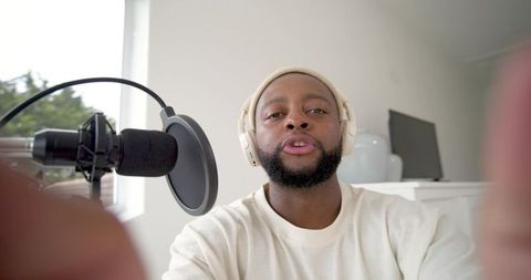 African american man recording podcast in bright home studio wearing headphones and beanie