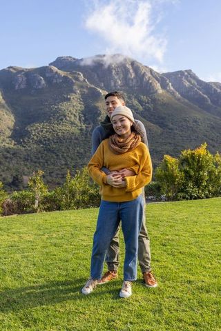 Asian couple embracing on mountain foothills lawn wearing cozy knit sweaters and beanie