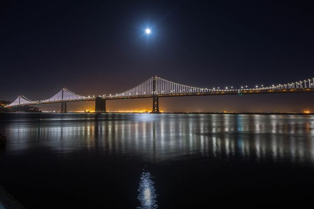 San Francisco-Oakland Bay Bridge lit up at night with city lights reflecting off the water and a bright full moon overhead. The image can be used for travel brochures, cityscape portfolios, or wallpaper backgrounds showcasing urban architecture and nighttime views.
