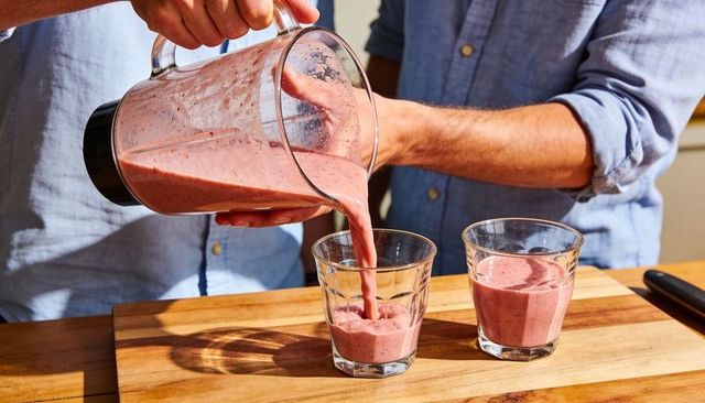 Couple pouring berry smoothies from blender pitcher into glass tumblers on wooden counter