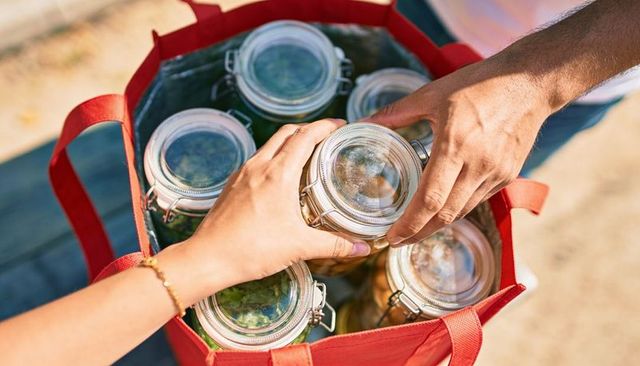 Hands exchanging glass jars in red tote bag filled with preserved food outdoors sunlit