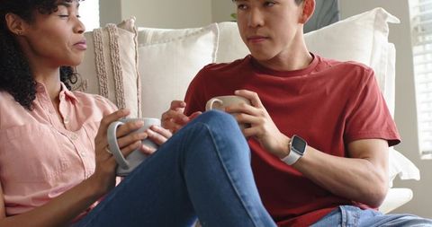 Diverse couple relaxing on sofa holding mugs, cozy conversation and home comfort