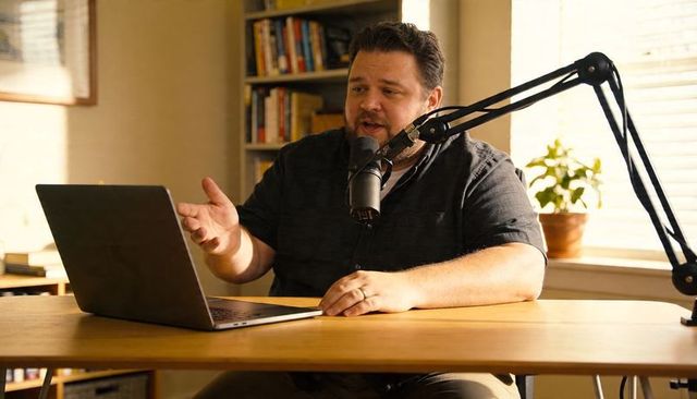 Man recording podcast in warm home office speaking into microphone with laptop