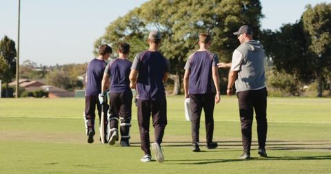 Diverse male cricket team walking off pitch with coach after training, carrying bats and pads