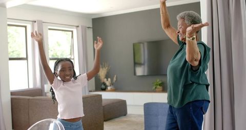 African american grandmother and granddaughter raising arms and smiling in living room