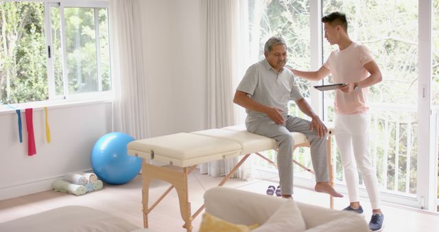 Physical therapist assisting senior man sitting on therapy table in bright, airy room. Ideal for use in healthcare, wellness, and elderly care promotions. Showcases rehabilitation and medical support for seniors.