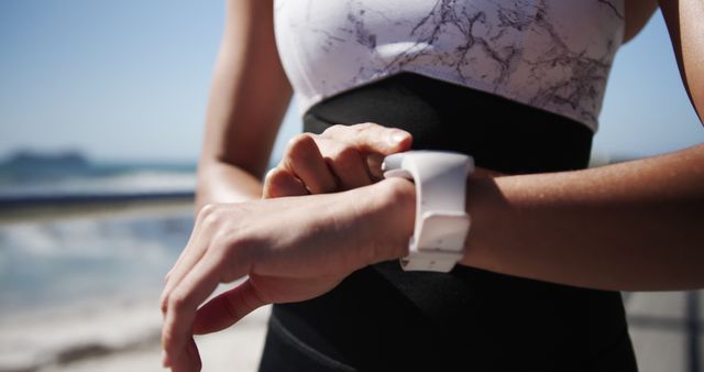 Athletic woman wearing sporty outfit engaged in monitoring heart rate on her smartwatch by the beach. Ideal for topics related to fitness, health, technology in sports, outdoor activities, or summer workouts. Useful for illustrating articles on fitness tracking, wearable technology, healthy lifestyle or exercise routines.