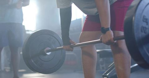 African male performing loaded barbell deadlift in gym with partner for strength training