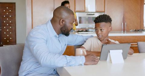 African American father mentoring son on tablet at modern kitchen table, family learning