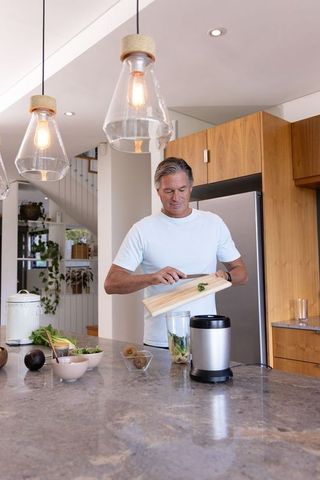 Middle-aged man preparing green smoothie in contemporary kitchen with wood cabinets