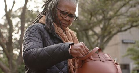 Senior african american woman searching handbag outdoors in quilted jacket and scarf