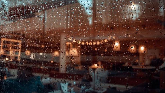 Rain-speckled window revealing warm restaurant interior with glowing pendant lights at dusk