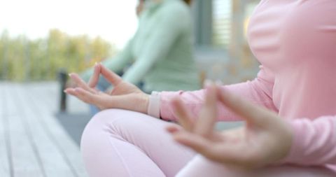 Female friends meditating on wooden balcony deck in pastel activewear with smartwatch