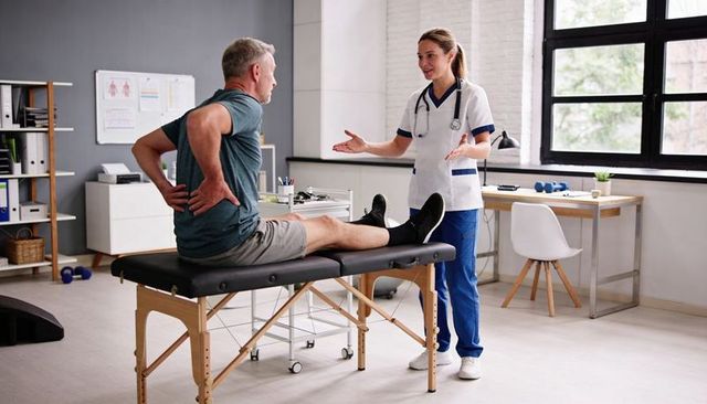 Middle-aged man holding lower back on treatment table consulting female clinician in rehab clinic