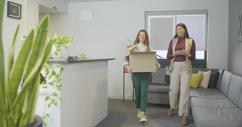 Smiling diverse coworkers walking into modern office carrying moving box, plant and smartphone