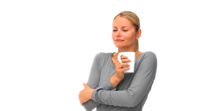 Blonde woman in grey sweater holding cup of coffee and appearing relaxed with closed eyes against light background. Ideal for advertisements highlighting relaxation, comfort, and enjoying a moment. Useful for lifestyle blogs, wellness content, and coffee-related promotions.