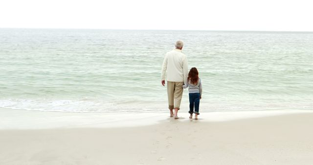 An older man and a young girl walk hand-in-hand at the beach. Their footprints mark the sandy shore as they face the calm ocean, suggesting a peaceful and bonding moment. Suitable for concepts of family bonding, generational relationships, tranquility, and leisure activities.