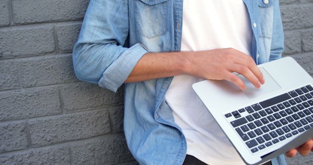Individual in denim shirt using laptop while leaning against gray brick wall. Suitable for themes related to remote work, modern technology, outdoor activities, and casual lifestyle.