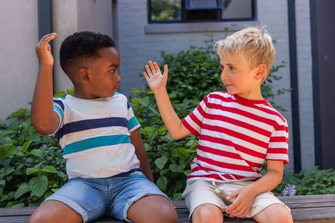 Two boys high-fiving on wooden bench in sunny courtyard wearing striped tees and shorts