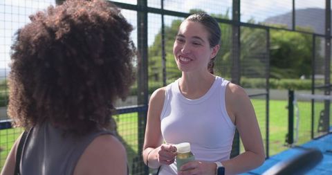 Diverse female friends chatting on sunny padel court holding water bottle
