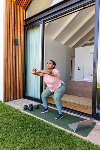 African american woman squatting on mat at doorway during outdoor workout with laptop