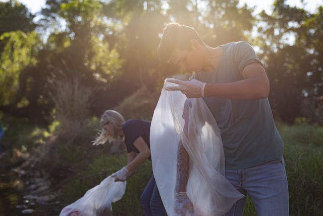 Male and female Caucasian conservation volunteers cleaning up river in the countryside, picking up rubbish holding rubbish bags. Ecology and social responsibility in rural environment.