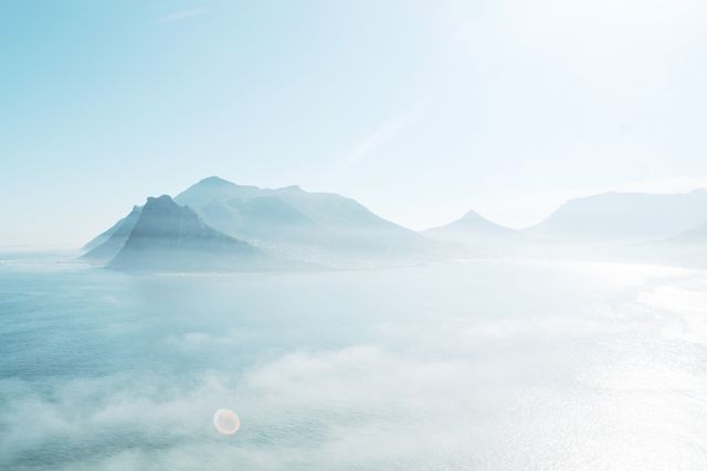Beautiful panoramic view of a coastal mountain range with mist rising from the water. Clouds gently hug the mountain peaks, creating a serene and tranquil atmosphere. Ideal for use in travel brochures, nature websites, relaxation and meditation materials, or as a background image for presentations emphasising calmness and peace.