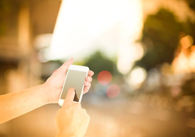 Hands interacting with a smartphone outdoors with a blurred street in the background. Ideal for illustrating mobile communication, technology, urban lifestyle, social media usage or modern connectivity themes in advertisements, blogs, or social media content.