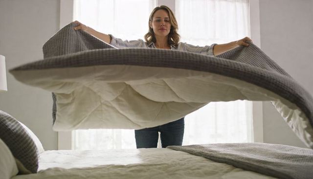 Woman straightening gray waffle comforter over cream duvet in bright minimal bedroom morning routine