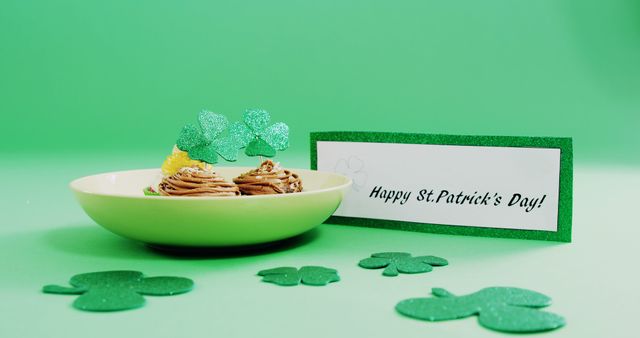 Cupcakes decorated for St. Patrick's Day placed on a green background with a greeting card and shamrock cutouts. Ideal for use in holiday marketing, social media promotions, and festive event invitations.
