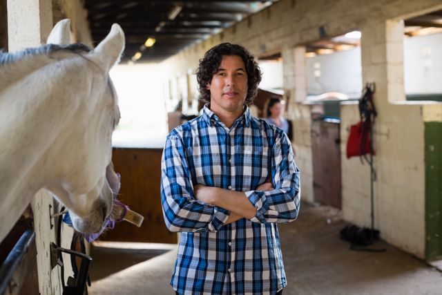 Man standing confidently with arms crossed in a ranch stable, wearing a plaid shirt. Horse visible in the foreground, suggesting a rural or agricultural setting. Ideal for use in articles or advertisements related to farming, ranching, equestrian activities, or rural lifestyles.
