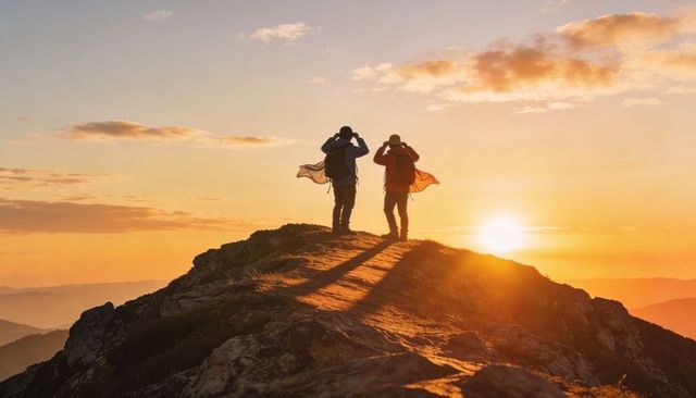 Two hikers watching golden hour sunrise from rocky mountain summit silhouetted panorama