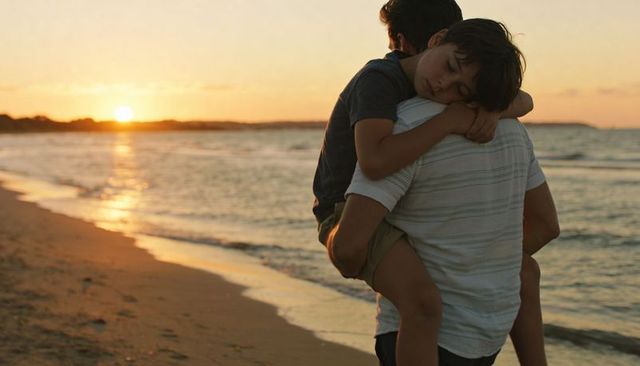 Father carrying sleeping son piggyback walking along shoreline at golden ocean sunset