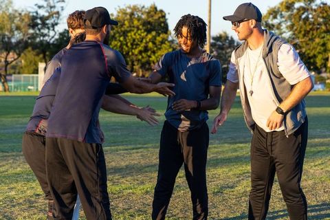 Diverse male teammates huddling with coach on sunlit park field promoting teamwork
